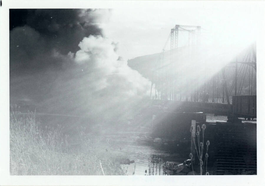 A photograph of Bridge 50.1 on the Camas Prairie Railroad next to the Kamiah Mine. Lens flare obscures most of the photo, but smoke can be seen billowing from a fire on the railroad bridge.