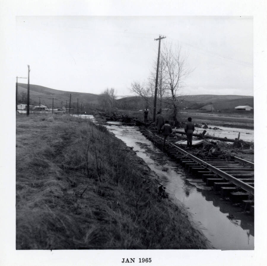 A photograph of ice jammed railroad tracks on the Camas Prairie Railroad near Lapwai Creek. There is debris on the tracks, as well as several people standing on the tracks, which are surrounded by icy water.