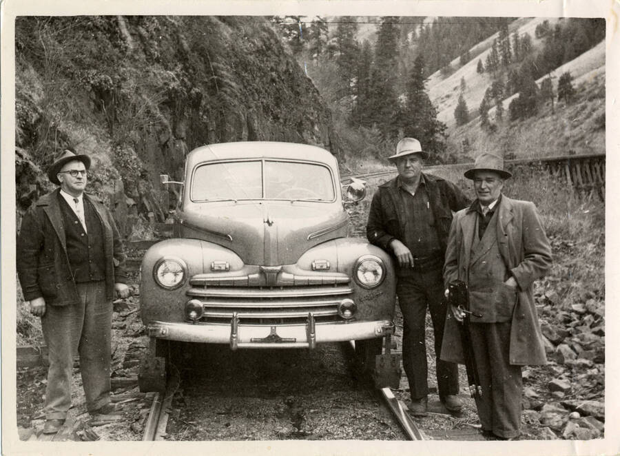 A photograph of a stationary passenger automobile. Three unidentified men pose around the car.