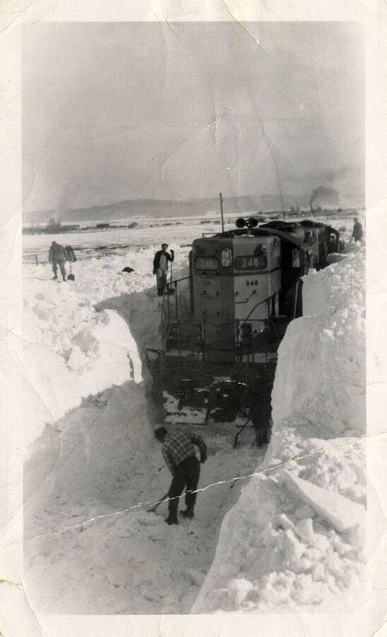 A photograph of Union Pacific Engine 248 stuck in its place on the tracks near Grangeville because of the immense amounts of snow. There are several workers in the photograph that are working to dig the train out.