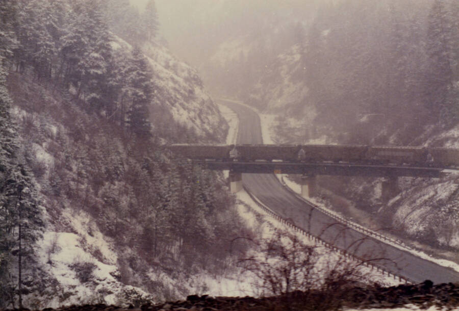 A color photograph of a freight train going over a bridge in a snowy wooded area.