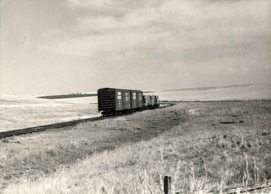 A photograph of two 40-ft box cars dwarfing the three G.E. diesel units enroute to Craigmont, Idaho on the Nez Perce Railroad. Through the shallow valley the light rails carry the bountiful wheat harvest of the rich Camas Prairie region.
