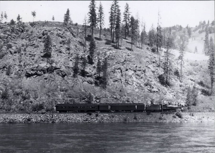 A photograph of a train moving alongside a river with several rolling hills adorned with trees and jagged rocks in the background.