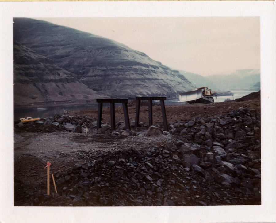 A photograph of the beginning piles of a bridge across a specific canyon. A construction vehicle sits alongside the river in the background. Mountains stretch past the river in the background.