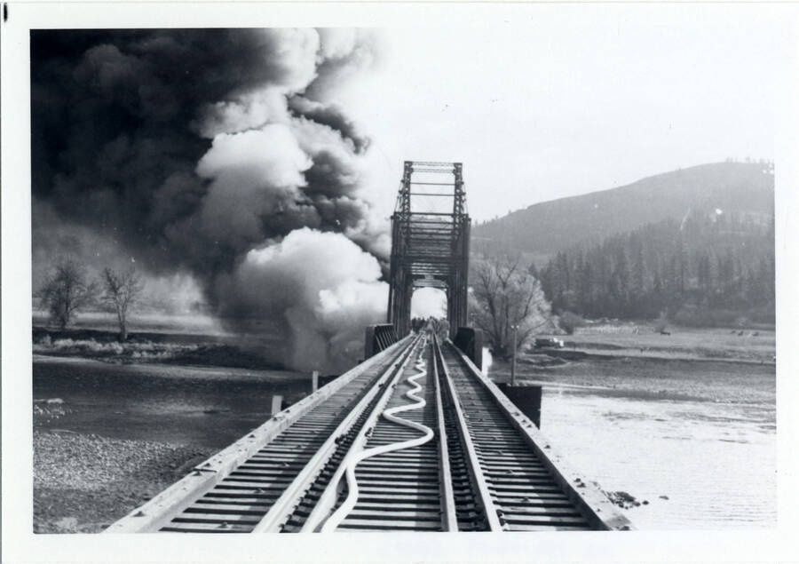 A photograph of Bridge 50.1 on the Camas Prairie Railroad next to the Kamiah Mine. Smoke billows from a fire on the railroad bridge. People can be seen on the bridge walking toward the fire.