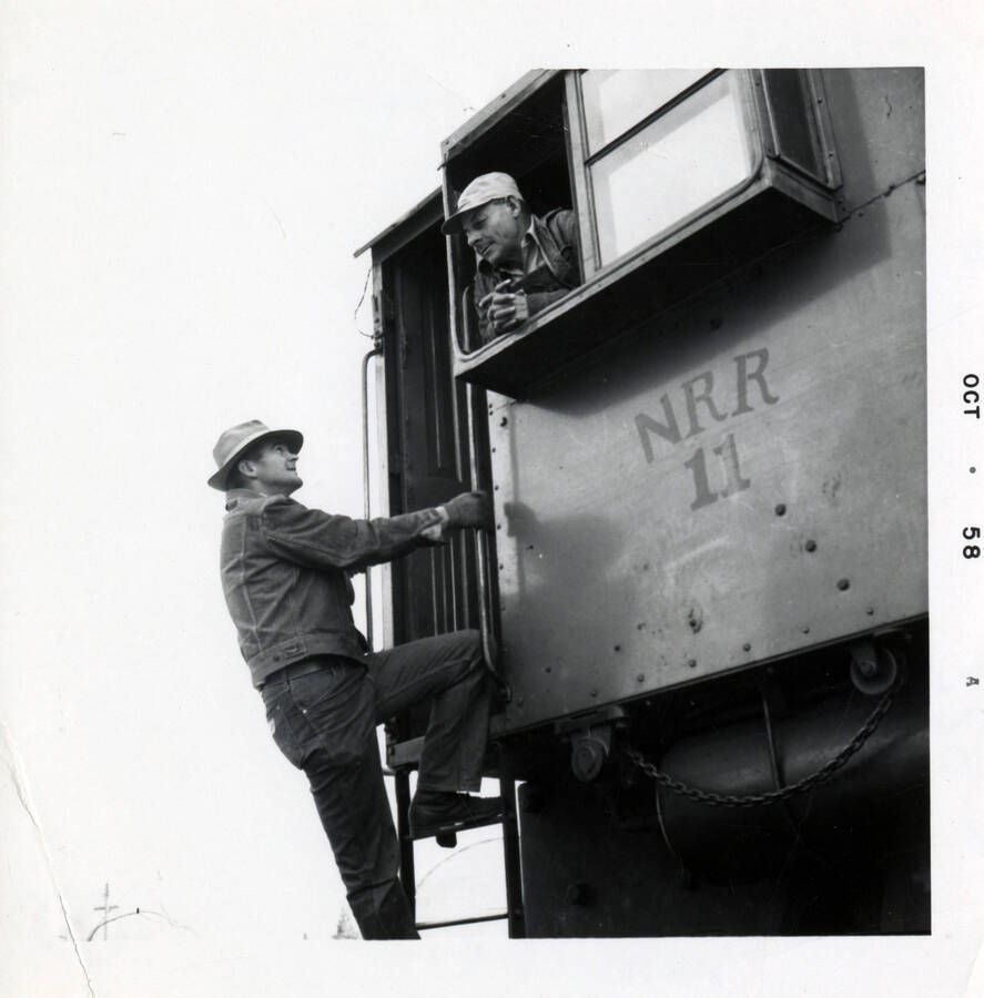 A photograph of a railroad worker climbing up a car to reach the cabin. Another worker looks out from a window in the cabin.