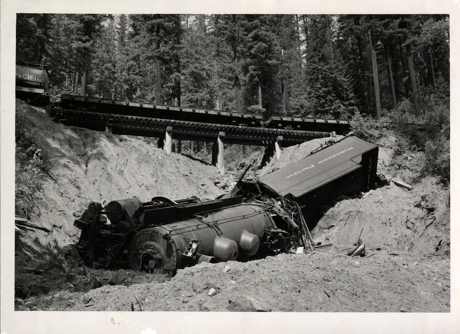 A photograph of derailed Northern Pacific Train Engine 1648. 'NP 1648 Pulling Camas Prairie Railroad Company Logger train hit a soft spot in track at MP 22 near Poorman Idaho June 1954. Jim Pilik hurt and fireman also. Engine cut up for scrap on the spot.' 'NP #1648 was going up to Headquarters branch and they hit a slip-off and the locomotive runned over on the left side and slid down into a hole.'