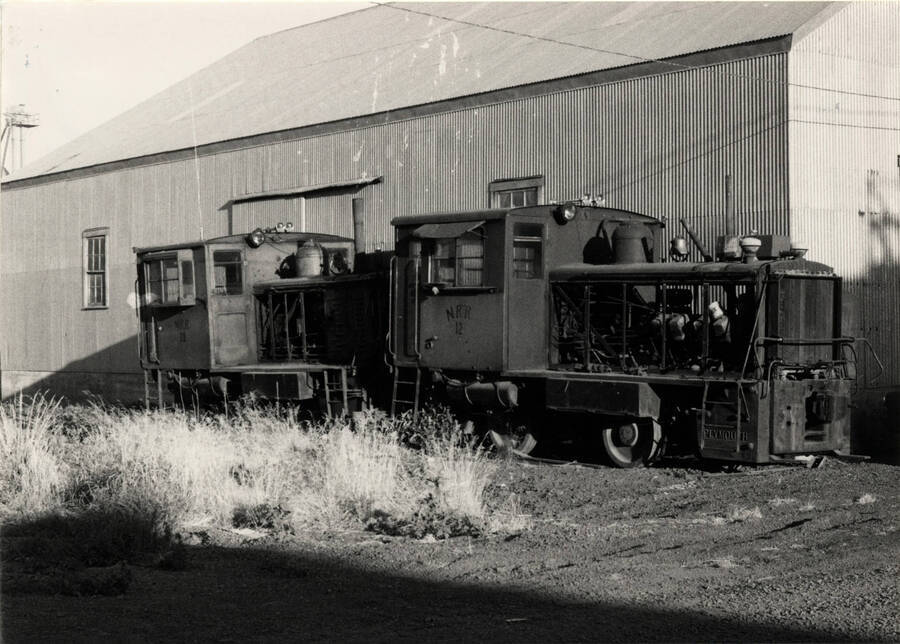 A photograph of Train Engines No. 11 and 12, of which wait out an uncertain future in Nez Perce, Idaho in the summer of 1973. No. 11, a 1936 built Plymouth, 250 HP was acquired from the Ohio & Morenci in 1952. No. 12, a 1935 built Packard, 190 HP was acquired from the Detroit Harbor Terminals in 1952, at a bargain price of $40,000/00 for the twon engines. The two gas powered locomotives pulled the Nez Perce out of the Depression, using only $5.00 worth of gasoline for the 27 mile round trip to Craigmont, Idaho, where a steam locomotive would burn $30.00 worth of coal. For 20 years, the tiny four-wheelers pulled the heave wheat loaded box cars out of the Nez Perce prairie.