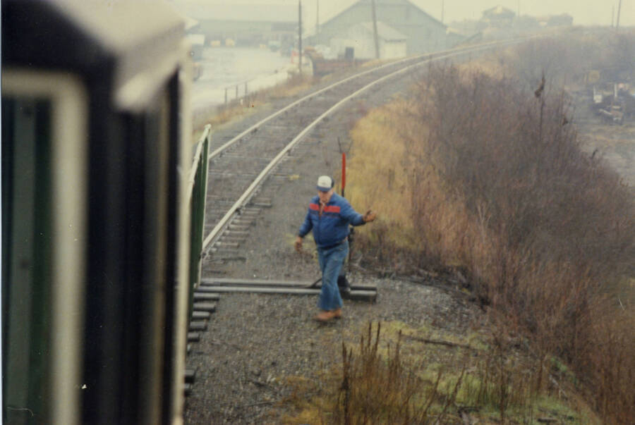 A color photograph of a man standing near the train tracks pictured from the cab of a train.