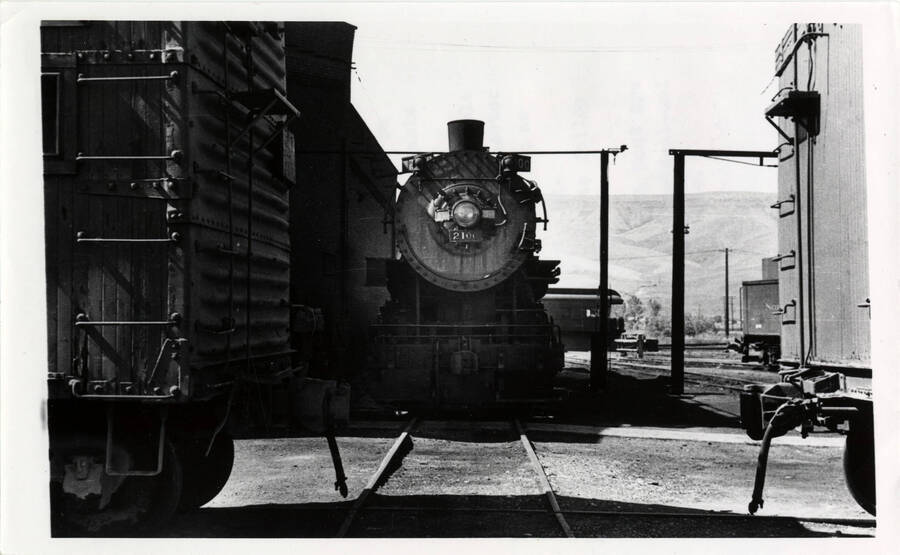 A photograph of the front-facing view of a stationary locomotive engine in Lewiston Train Yards. It appears as if the train is headed straight toward you. 'UP 2-3-2 #2100 (name of engine) sits next to the roundhouse. The private car of NP Idaho Division superintendent B.L. Slorah is on the turntable in the background.'