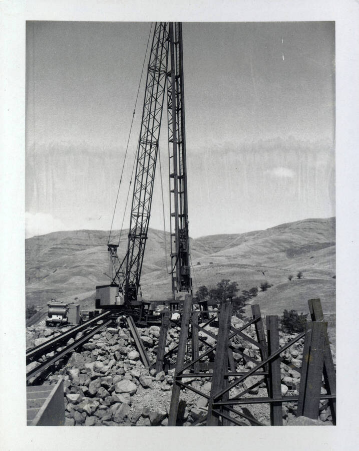 A photograph of a partially constructed Wawawai Bridge. A metal crane maintains the focal point of the picture. Rolling hills extend in the background.