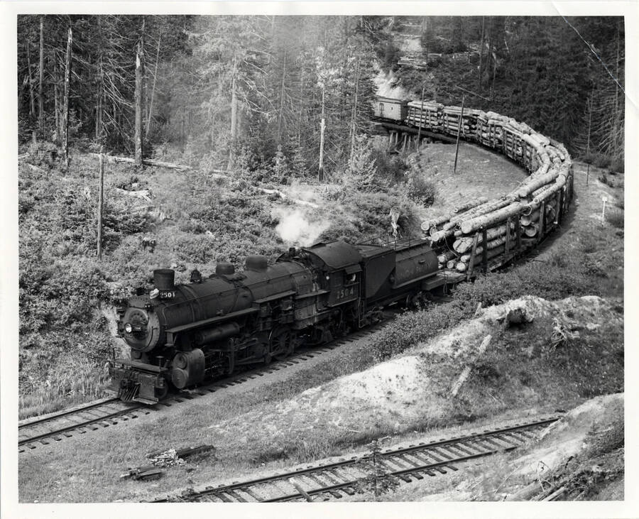 A photograph of train engine #2504 carrying a freight of logs across the mountainside out of Headquarters, passing a logging spur of Potlatch Forest Industries.