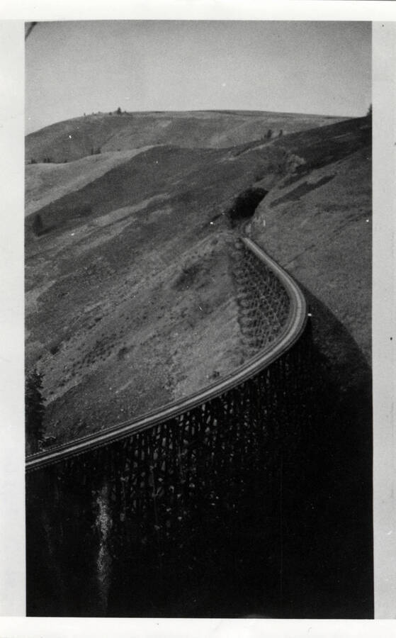 A photograph of an unidentified railroad bridge, leading into a tunnel. Rolling hills extend in the background.