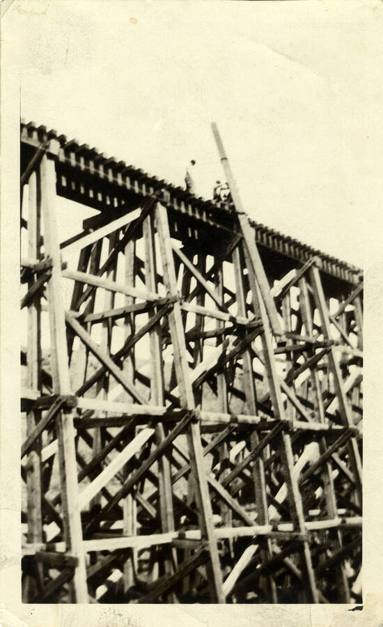 A photograph of several construction workers standing atop a bridge, pulling a train engine out of a river. The train engine is not pictured in the photograph captured.