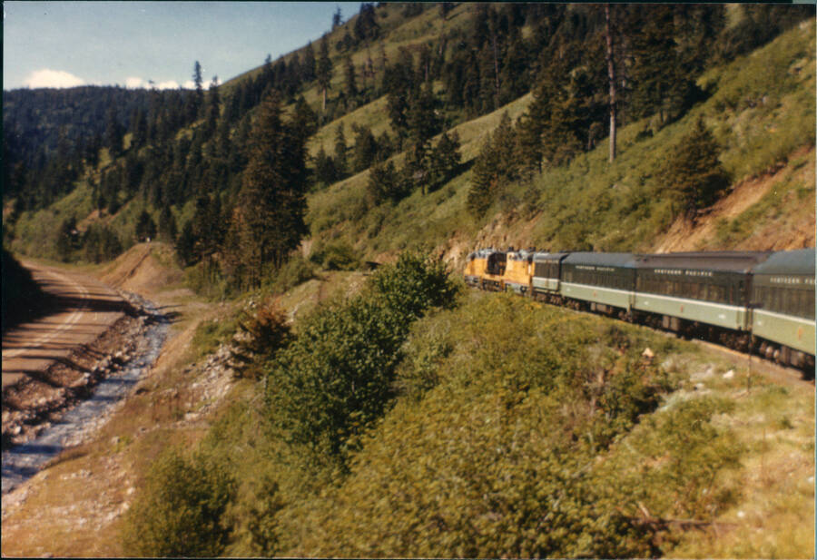 A freight train moves through mountainous terrain. There are sparse trees along the mountainside in the left-side of the picture. A river runs to the left and there is a road to the left of the river.