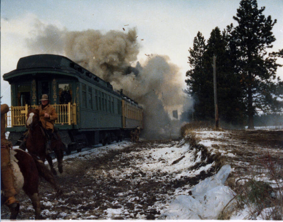 A photograph of a train stopped at the Craigmont Junction on the set of the film "Breakheart Pass". Description from Bill Clem: "Black powder smoke bombs resulting from dynamite being thrown into supply car as Indians ransacked car to secure rifles and ammunition. 'Dynamite' was thrown by Deakin (Charles Bronson) riding by on horseback (rider in picture is not Bronson)."