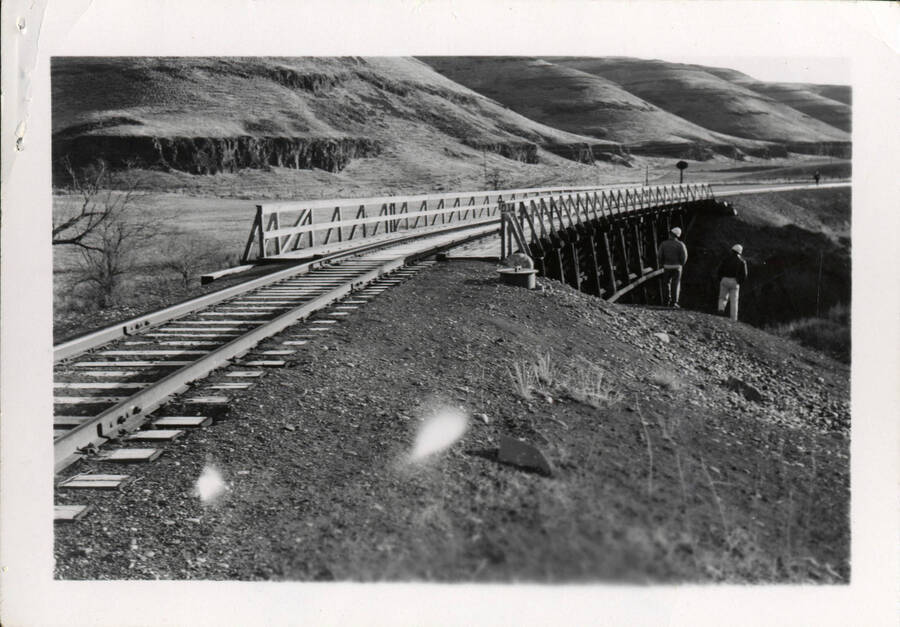A photograph of the Riparie Bridge extending across a ravine in Walla Walla. There are a few men wearing construction hats standing to the right of the bridge, with rolling hills extending in the background.