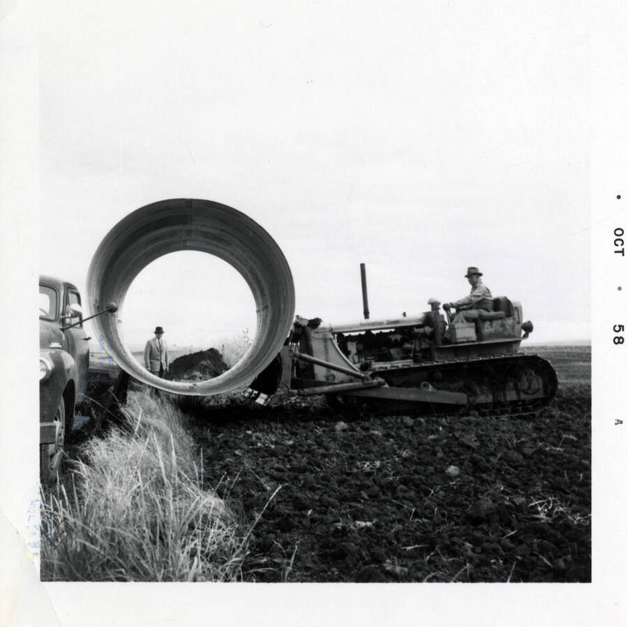 A photograph of a worker riding a tractor while moving what appears to be a large piece of pipe.