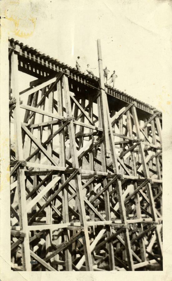 A photograph of several construction workers standing atop a bridge, pulling a train engine out of a river. The train engine is not pictured in the photograph captured.