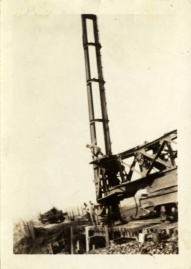 A photograph of pile driving occurring on bridge 66 of the Camas Prairie Railroad. Two men stand on the railroad scaffolding while another is atop the pile driver machinery.