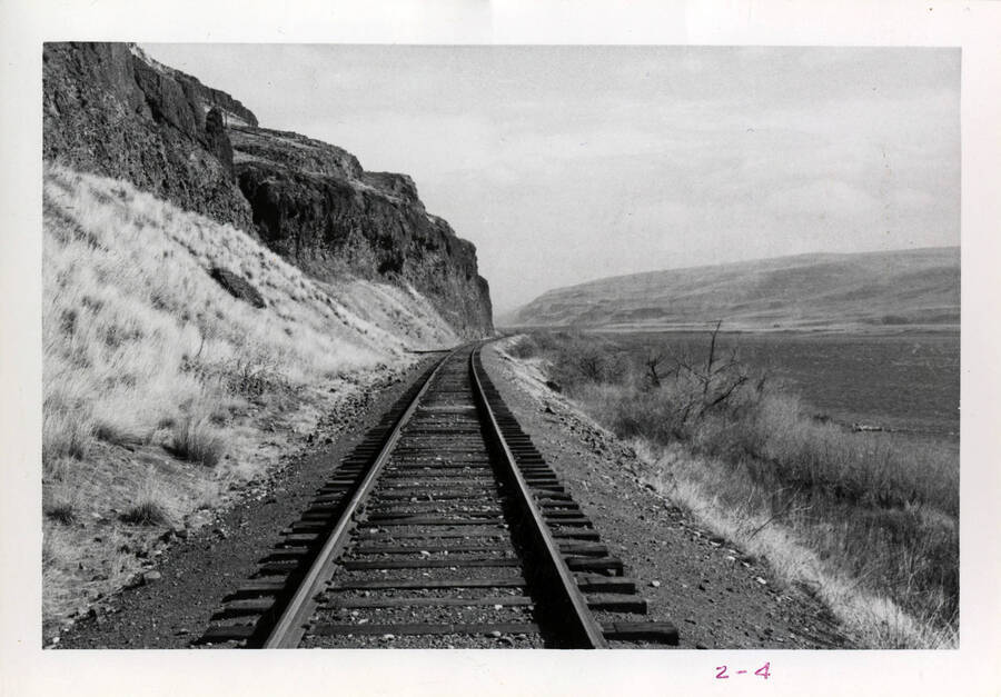 A photograph of the Snake River Valley Railroad, extending far into the background. The Snake River bends along to the right of the railroad, with rolling hills across the water, and cliffs and steep rock fixtures to the left of the railroad.