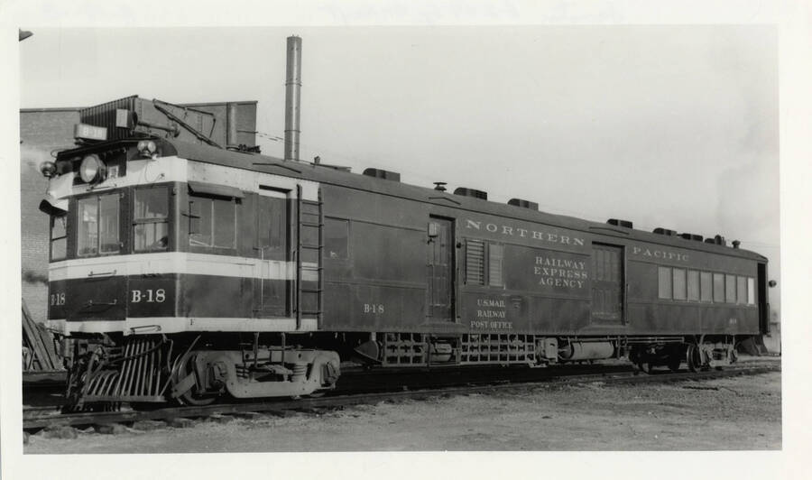 Northern Pacific B-18 railway post office locomotive stopped on railroad tracks next to a brick building.