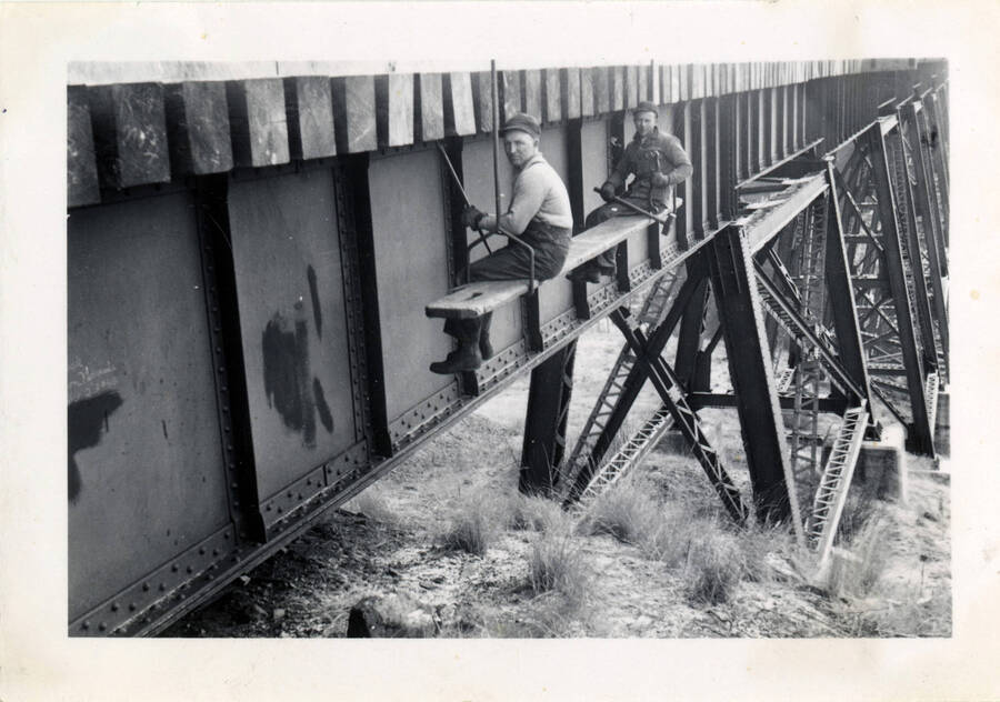 A photograph of construction workers taking a break while sitting on a suspended metal bar intended for the construction of bridge 38 of the Camas Prairie Railroad.