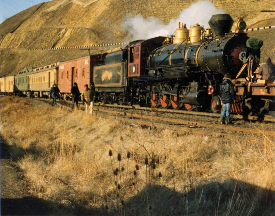 A photograph of a train stopped at the Arrow Junction during the filming of "Breakheart Pass". Description from Bill Clem: "Taken at Arrow Junction on maiden trip east from Lewiston to Orofino, Idaho. At this point diesel unit and road caboose were detached and run ahead of "Breakheart Pass" train so that helicopter shots could be made of steam locomotive pulling vintage cars along the Clearwater river. Although tender is stacked with cord wood, steam was made by hand firing locomotive with coal."