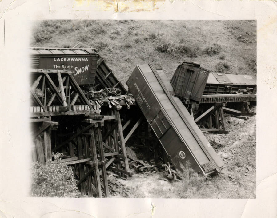 A photograph of what appears to be a large train accident, with several train cars toppling over the edge of the tracks onto the rolling hills. The train engine isn't pictured, only the freight cars that have been affected.