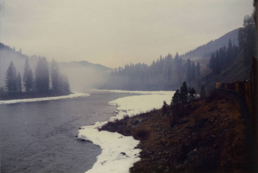 A color photograph of a river just off the bank of train tracks pictured from the cab of a freight train.
