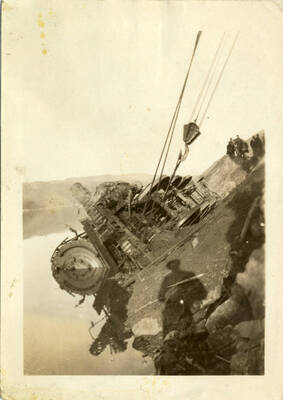 A photograph of a toppled, partly submerged train engine being raised out of the water using construction machinery. Captured in the background is a group of several people watching the process happen.