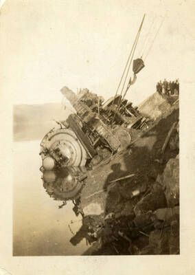 A photograph of a toppled, partly submerged train engine being raised out of the water using construction machinery. Captured in the background is a group of several people watching the process happen.