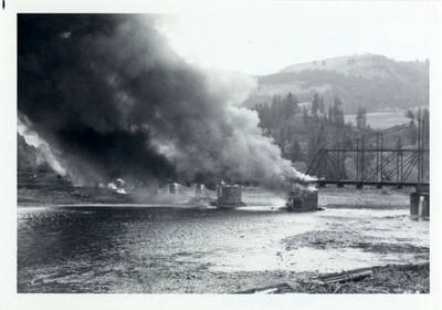 A photograph of Bridge 50.1 on the Camas Prairie Railroad next to the Kamiah Mine. Smoke billows from a fire on the railroad bridge. People can be seen on the bridge walking toward the fire.