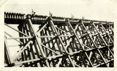 A photograph of several construction workers standing atop a bridge, pulling a train engine out of a river. The train engine is not pictured in the photograph captured.