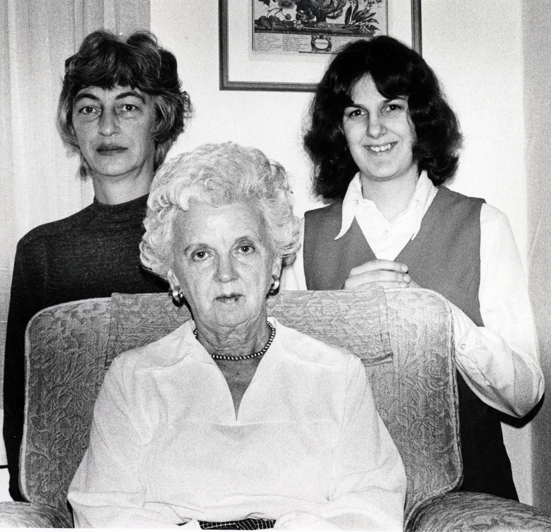 Photograph of Etta Mae Egland seated, looking at the camera, with two women standing behind Egland.
