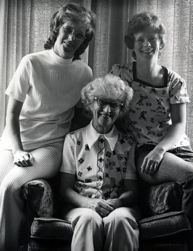 Photograph of Norma June Ohlson seated, smiling at the camera, with two women standing behind Ohlson.