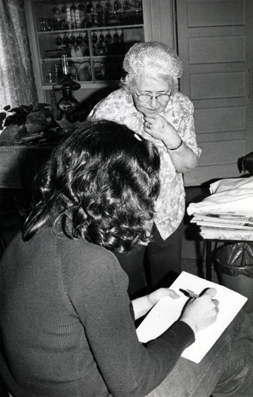 Photograph of a seated woman with her back to the camera, writing, with Nellie Smith looking on.