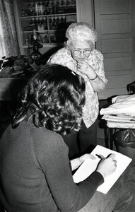 Photograph of a seated woman with her back to the camera, writing, with Nellie Smith looking on.