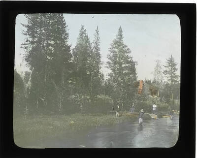 Glass lantern slide of a camp site with approximately four men standing near the river and a cabin in the background.