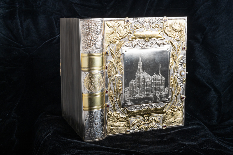 Jewelry box is sitting on bottom edge as if it is a book. Edge is styled as a book spine, featuring a silver relief of pine cones, the Idaho state seal (the only state seal designed by a woman), and the dates 1863 (the year Idaho Territory was created) and 1893 (the year of the exposition). Front cover features etching of original Administration Building, gold reliefs of wheat shafts and representations of industries that were important to early Idaho's development.  