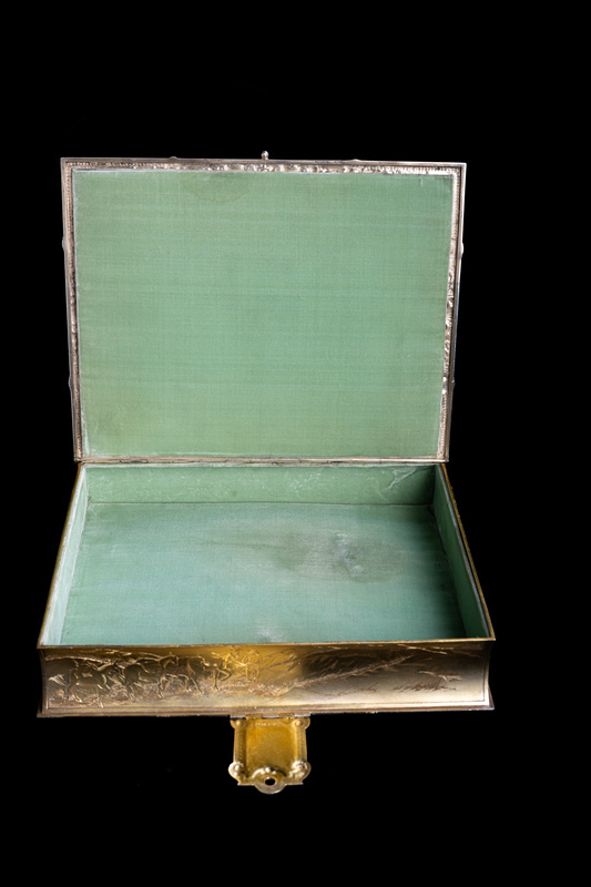 Jewelry box is open to reveal green velvet interior. Long edge golden relief of scene of miner with pack mule train is visible, as is the underside of the box clasp. 