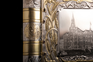 Close up detail of one long edge of object which appears as the spine of the book. Featuring a golden relief rendering of the state seal of Idaho. The state seal was designed by Emma Edwards Green, the only women to design a state seal in America. The seal includes imagery meant to evoke the foundational characteristics of Idaho. 