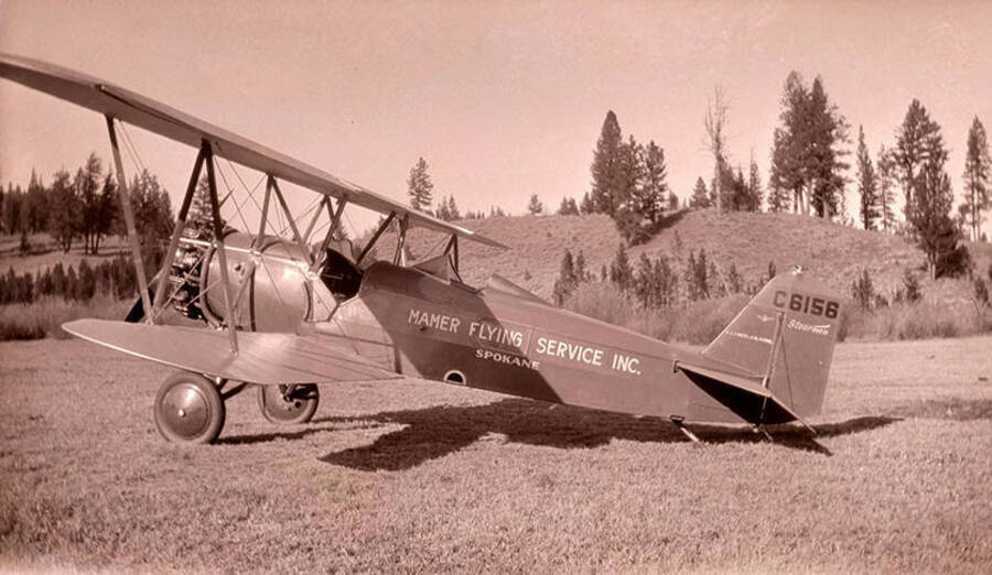 An airplane with a banner that reads: 'Mamer Flying Service, Inc.' parked in a meadow near the Stonebraker Ranch. Pilot Nick Mamer flew the plane to the Chamberlain Basin from Spokane, Washington. The plane is numbered 'C6156, Stearman.'
