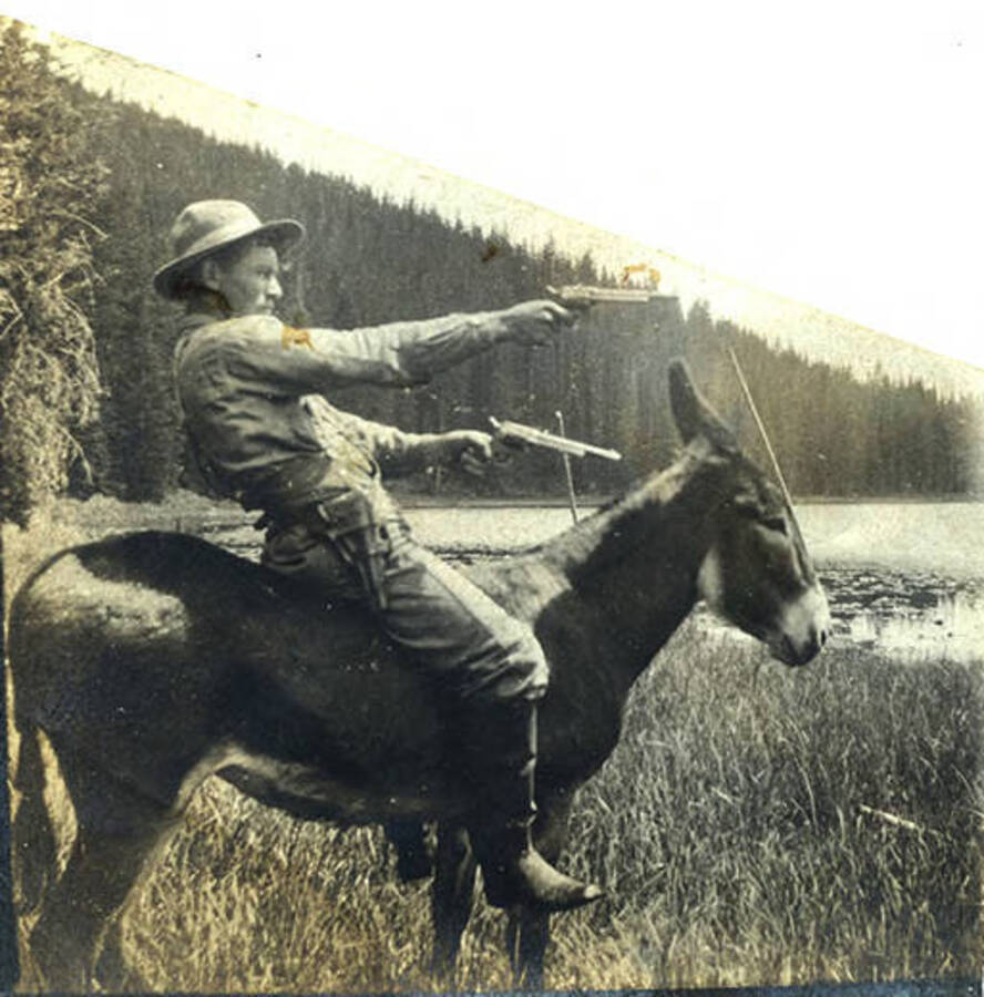 Sumner Stonebreaker sits on Bosco the mule holding two pistols near the river. The photo captions reads: 'Wild Bill. Throw up your hands!'