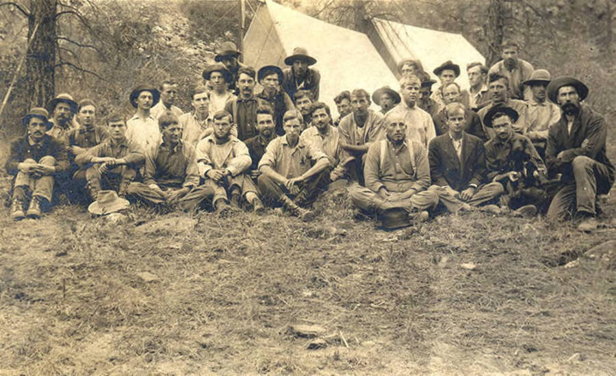 Group of men sitting for a photograph with tents in the background.