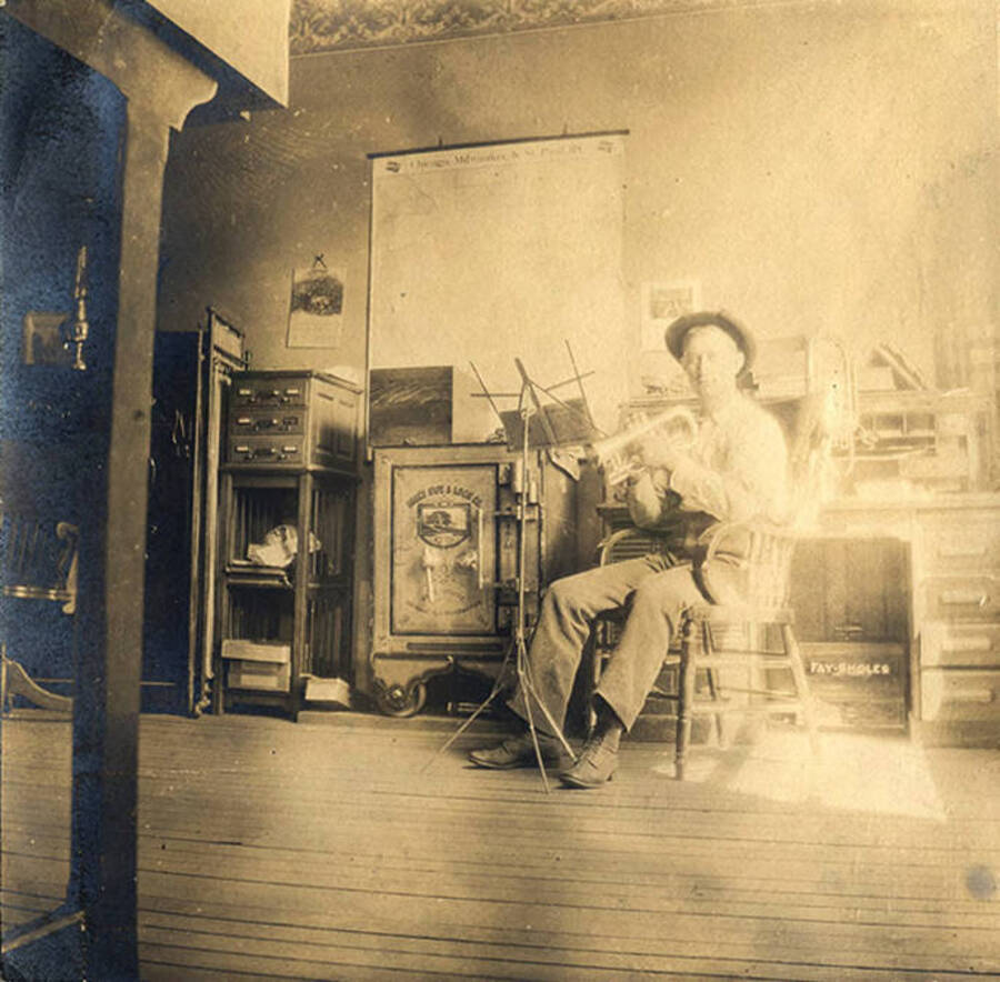 Photo caption reads: 'Teddy, assistant cashier of the Bank of Stites.' Teddy poses with his trumpet and music stand while sitting in a desk chair in the bank near the safe.