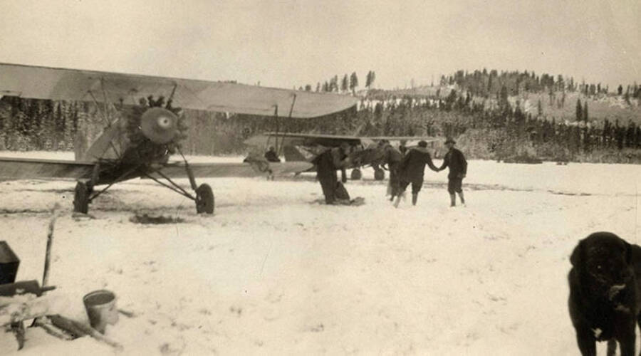Two Zenith airplanes parked in a snowy meadow near Stonebraker Ranch in Chamberlain Basin. A dog stands in corner with men shaking hands near the airplanes.