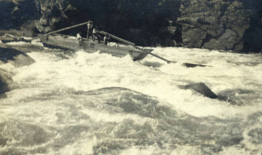 A boat with two sweeps rides the rapids on the South Fork of the Salmon River along a rocky edge.