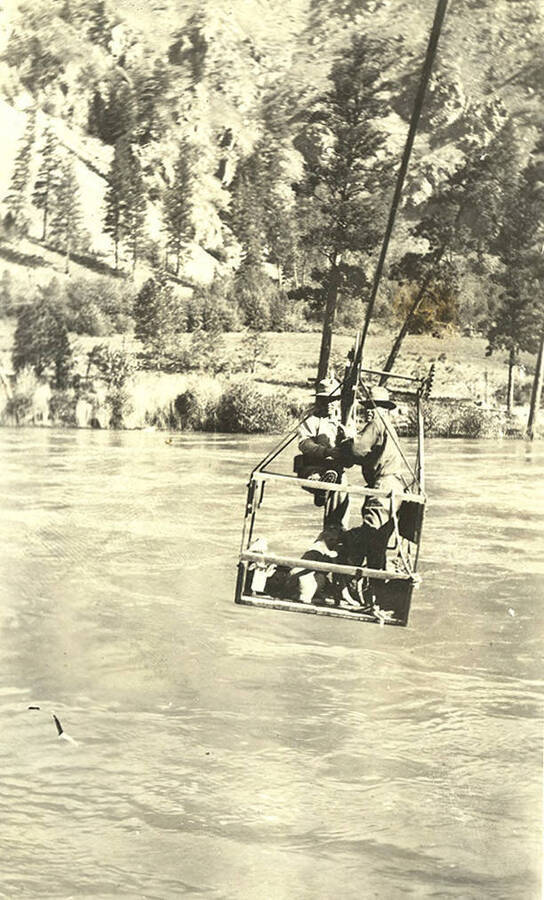 Two men use pulleys to move the cable car across the river. Photograph printed at the Owl Drug in Lewiston, Idaho.