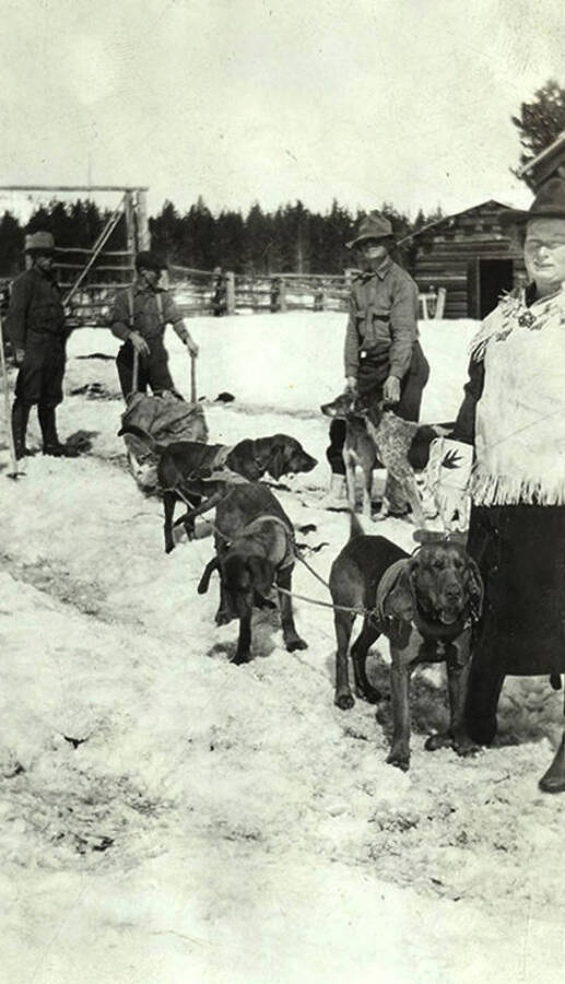 A dogsled team prepares for a trip. Men and women stand near dogs outside the corral.